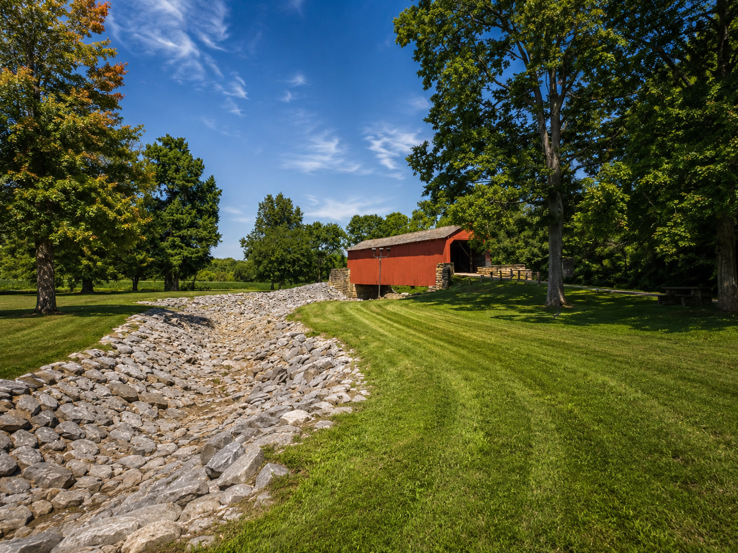 Wide scouting photograph of Mary’s River Covered Bridge in Illinois with blue sky, grass, trees and a rocky drainage channel leading toward the bridge.