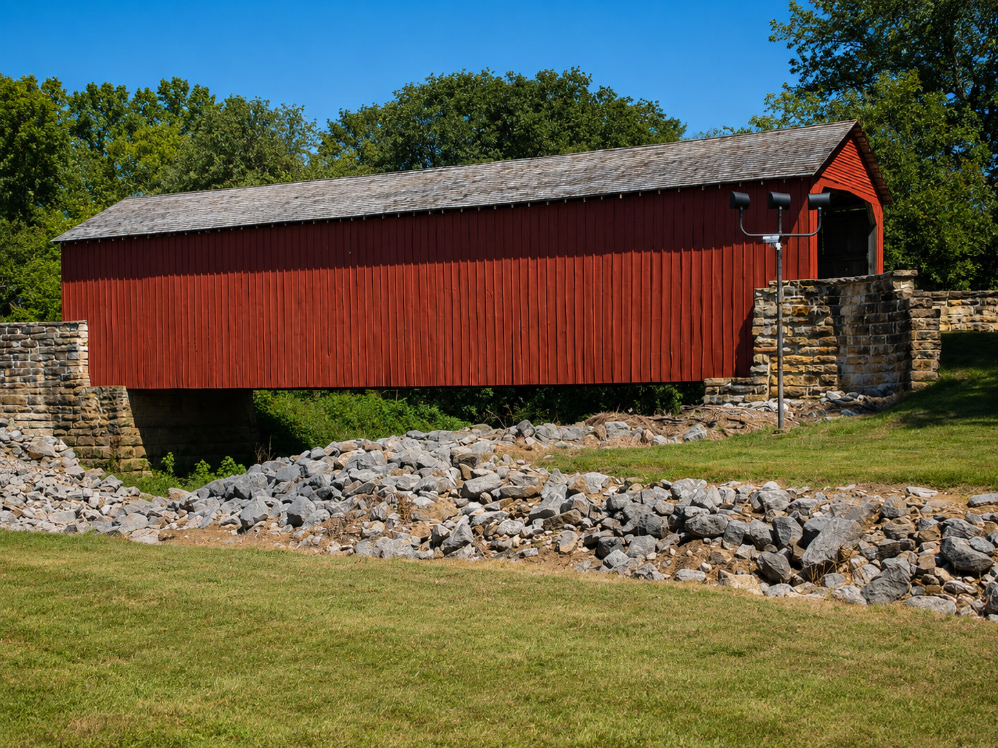 Closer view of a red covered bridge with stone supports, rocky drainage area and open grass in bright summer sunlight.