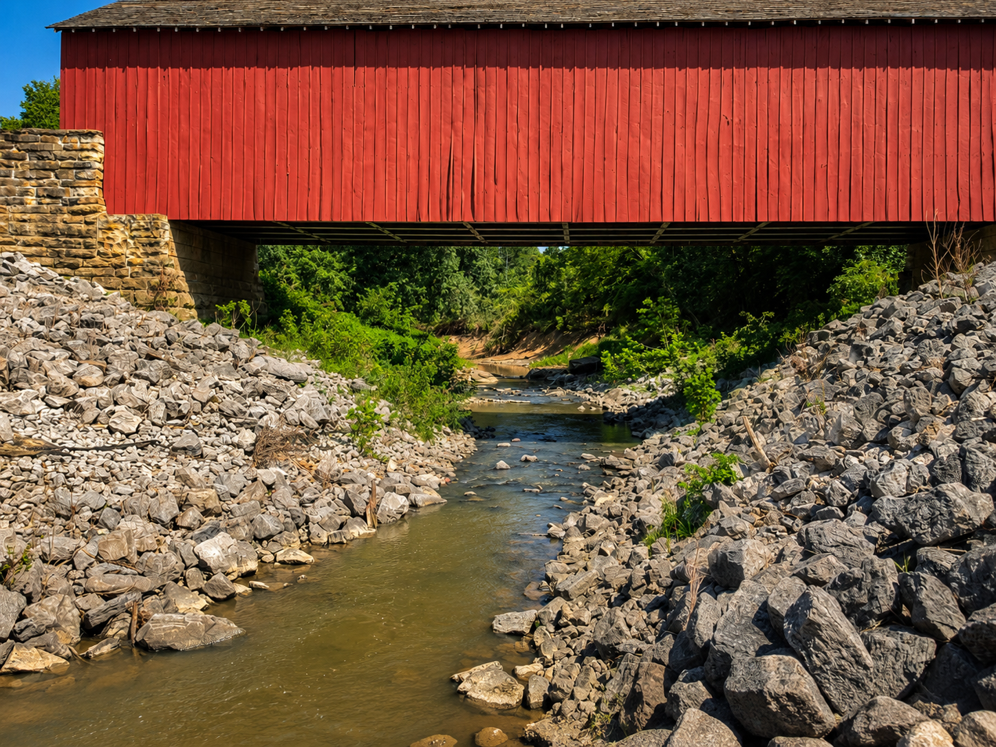 Tighter landscape composition of a creek flowing beneath a red covered bridge with rock banks framing both sides.