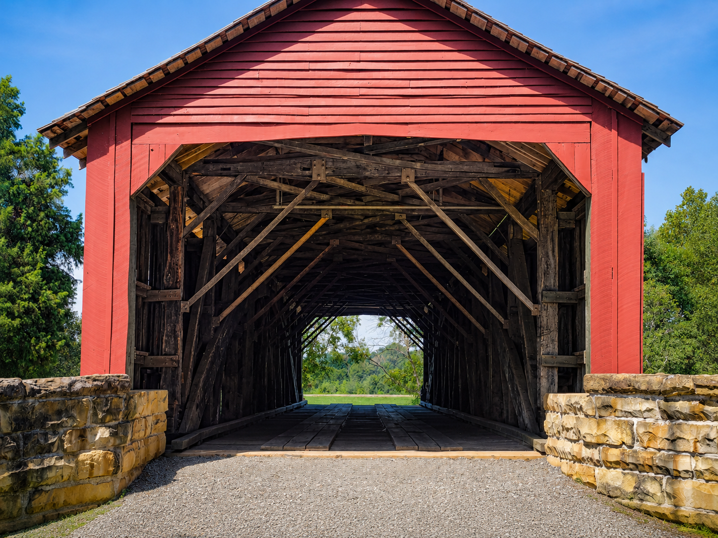 Straight-on view into the entrance of a red covered bridge with repeating wooden beams leading toward a bright opening at the far end.