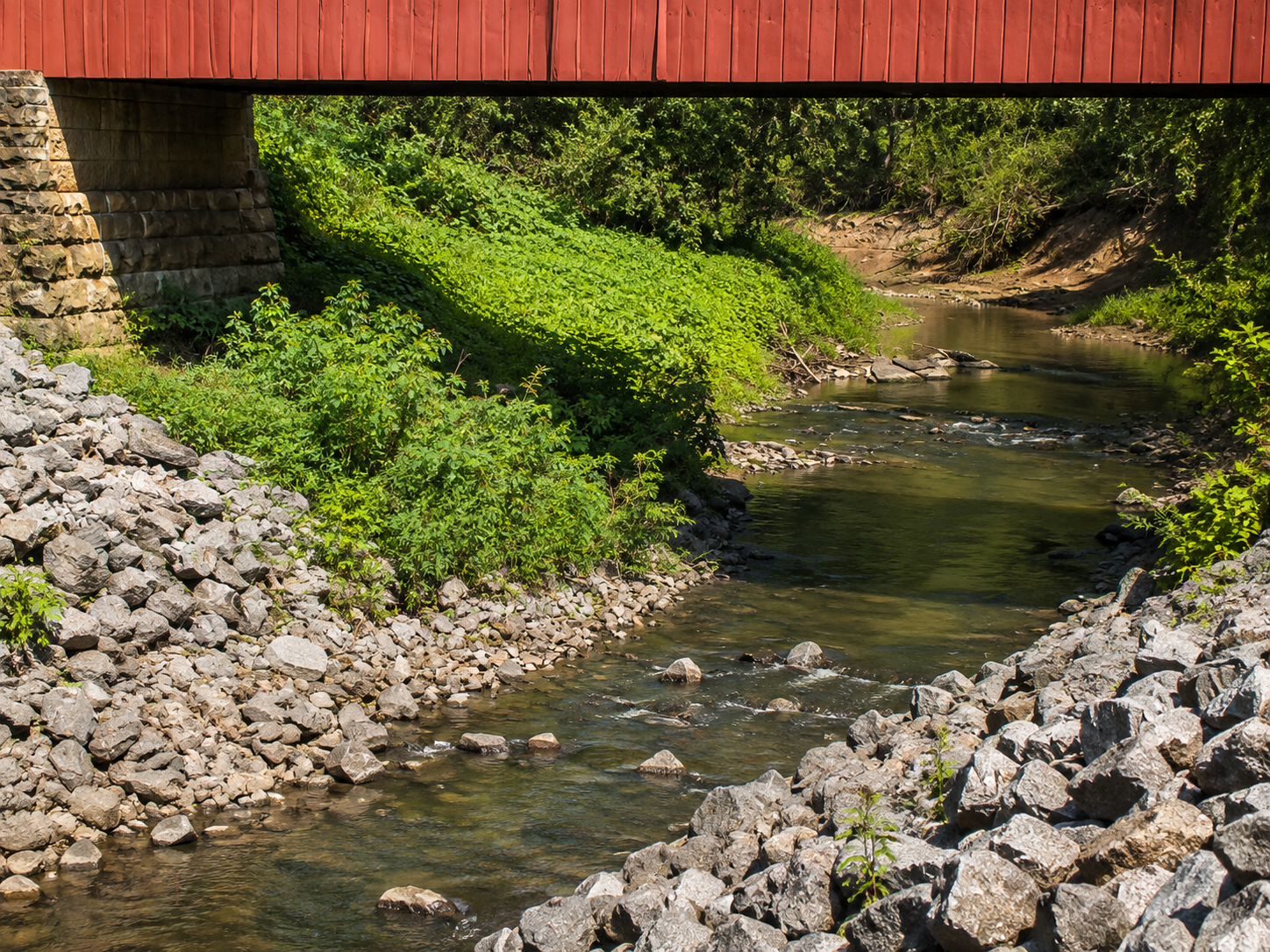 ight view of a shallow creek beneath a covered bridge, with rocks and green vegetation surrounding the water.