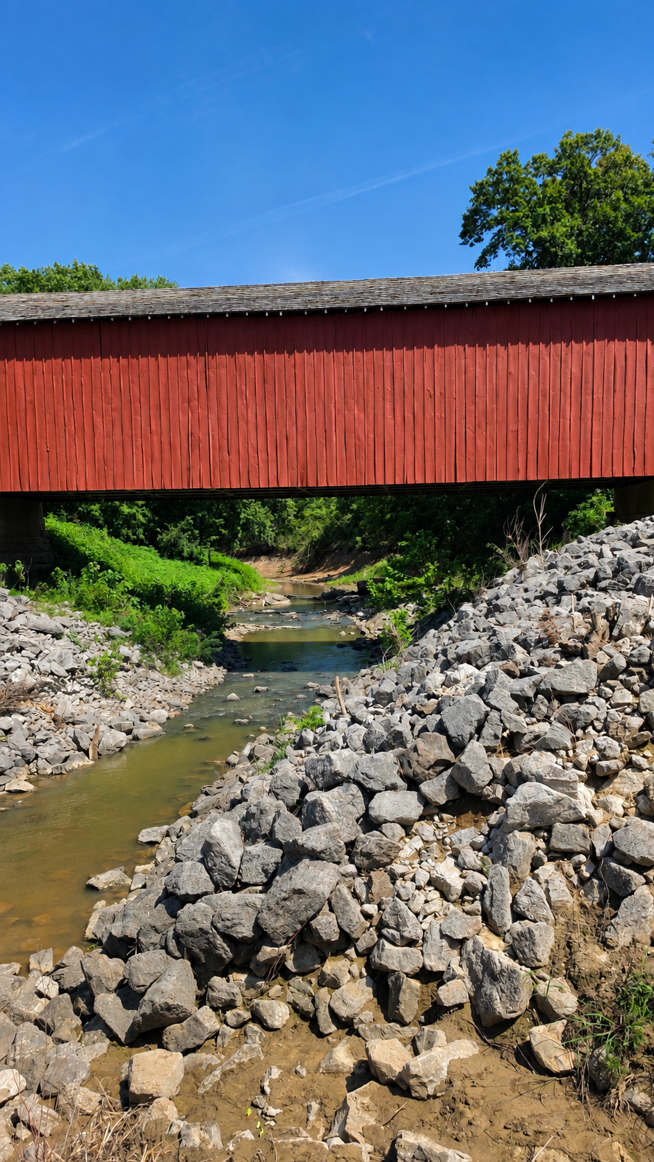 Vertical photograph of a shallow creek flowing beneath a red covered bridge, with a large rocky foreground and bright blue sky above.